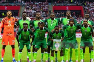 Nigeria national football team players pose for a group photo on the pitch, green kits with the goalkeeper in orange on the left, crowd in the stands behind them.