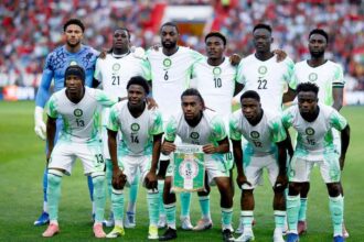 Nigeria national football team (Super Eagles) posing for a group photo on the field in white and green kits, with a banner held by the center player