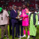 Soccer players and officials pose on a stadium field as a pink clad goalkeeper receives a trophy from an official in a suit  trybalnews