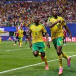 Two soccer players in yellow and green kits celebrate a goal on a green pitch as fans cheer in the stands behind them  trybalnews