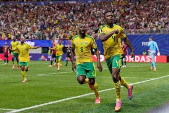 Two soccer players in yellow and green kits celebrate a goal on a green pitch as fans cheer in the stands behind them.