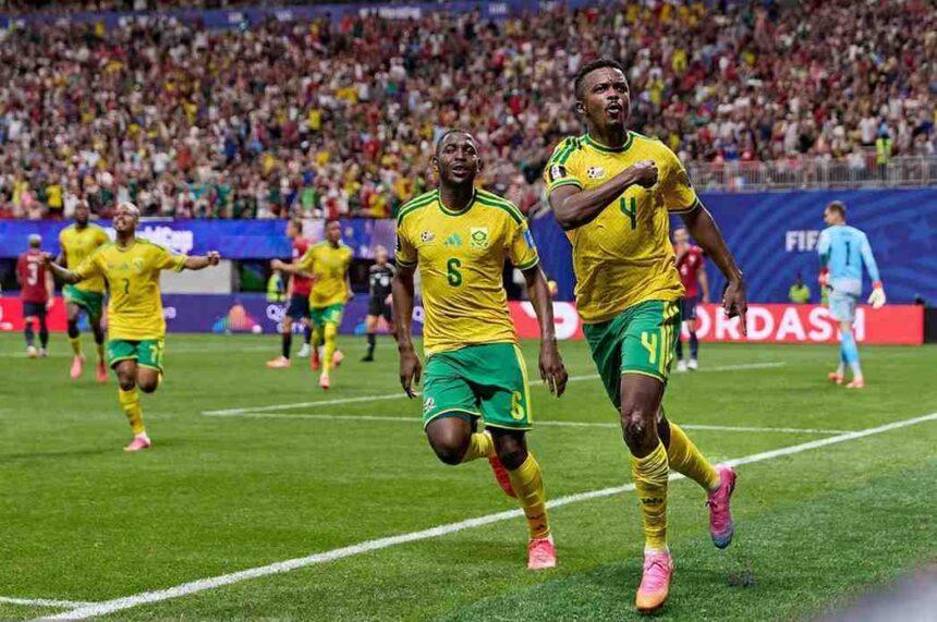 Two soccer players in yellow and green kits celebrate a goal on a green pitch as fans cheer in the stands behind them  trybalnews