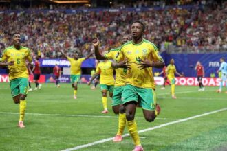 South African footballer Teboho Mokoena celebrates with arms raised after scoring a penalty kick. He wears a green and yellow South Africa jersey. The FIFA World Cup 2026 match between Czechia and South Africa is at Atlanta Stadium, June 18, 2026. Crowd and stadium visible in background.