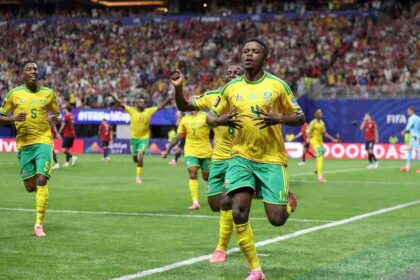 South African footballer Teboho Mokoena celebrates with arms raised after scoring a penalty kick. He wears a green and yellow South Africa jersey. The FIFA World Cup 2026 match between Czechia and South Africa is at Atlanta Stadium, June 18, 2026. Crowd and stadium visible in background.