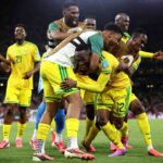 South Africa forward Thapelo Maseko 12 in yellow kit with green trim is mobbed by teammates after scoring He smiles while being hugged from behind by a player in white training vest South Africa 21 visible in background Stadium lights illuminate the night match on green pitch FIFA World Cup 2026 Group A match between South Africa and Korea Republic at Monterrey Stadium Guadalupe Mexico on June 24 2026 Team celebration shows emotion after first goal  trybalnews