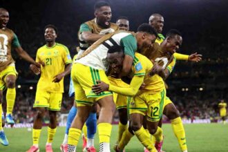 South Africa forward Thapelo Maseko #12 in yellow kit with green trim is mobbed by teammates after scoring. He smiles while being hugged from behind by a player in white training vest. South Africa #21 visible in background. Stadium lights illuminate the night match on green pitch. FIFA World Cup 2026 Group A match between South Africa and Korea Republic at Monterrey Stadium, Guadalupe, Mexico on June 24, 2026. Team celebration shows emotion after first goal.