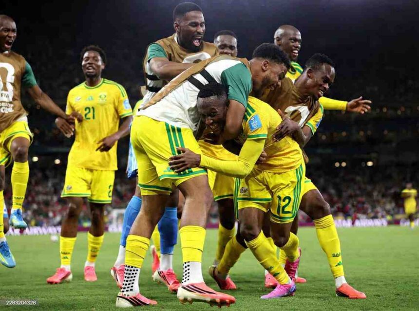South Africa forward Thapelo Maseko 12 in yellow kit with green trim is mobbed by teammates after scoring He smiles while being hugged from behind by a player in white training vest South Africa 21 visible in background Stadium lights illuminate the night match on green pitch FIFA World Cup 2026 Group A match between South Africa and Korea Republic at Monterrey Stadium Guadalupe Mexico on June 24 2026 Team celebration shows emotion after first goal  trybalnews
