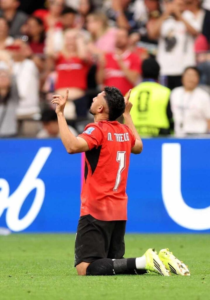 Egypt midfielder Mahmoud Trezeguet Hassan 7 kneels on the grass with his back to camera both index fingers pointing skyward in celebration He wears a red Egypt jersey with 7 and black shorts neon yellow boots visible The BC Place crowd in red fills the background during the FIFA World Cup 2026 Group G match vs New Zealand on June 21 2026  trybalnews