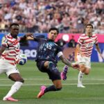 Soccer players clash for the ball in a match a navy kit player no15 kicks while red and white striped opponents approach with a crowded stadium in the background  trybalnews