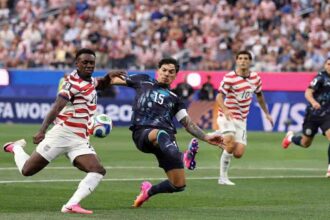 Soccer players clash for the ball in a match; a navy kit player (no.15) kicks while red-and-white striped opponents approach, with a crowded stadium in the background.