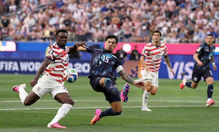 Soccer players clash for the ball in a match a navy kit player no15 kicks while red and white striped opponents approach with a crowded stadium in the background  trybalnews