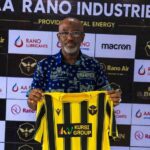 Usman AbdAllah a bald man with glasses and a gray beard wearing a blue patterned shirt holds up a Rancher Bees yellow and black vertical striped football jersey with KURSI GROUP sponsor logo He stands in front of a branded backdrop with AA RANO INDUSTRIES RANO LUBRICANTS Rano Air macron and Rancher Bees hexagon bee logo  trybalnews