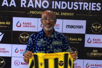 Usman Abd'Allah, a bald man with glasses and a gray beard wearing a blue patterned shirt, holds up a Rancher Bees yellow and black vertical striped football jersey with "KURSI GROUP" sponsor logo. He stands in front of a branded backdrop with "AA RANO INDUSTRIES", "RANO LUBRICANTS", "Rano Air", "macron", and Rancher Bees hexagon bee logo.