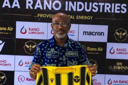 Usman Abd'Allah, a bald man with glasses and a gray beard wearing a blue patterned shirt, holds up a Rancher Bees yellow and black vertical striped football jersey with "KURSI GROUP" sponsor logo. He stands in front of a branded backdrop with "AA RANO INDUSTRIES", "RANO LUBRICANTS", "Rano Air", "macron", and Rancher Bees hexagon bee logo.