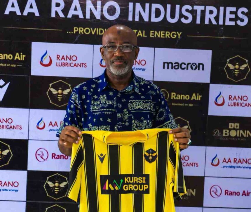 Usman AbdAllah a bald man with glasses and a gray beard wearing a blue patterned shirt holds up a Rancher Bees yellow and black vertical striped football jersey with KURSI GROUP sponsor logo He stands in front of a branded backdrop with AA RANO INDUSTRIES RANO LUBRICANTS Rano Air macron and Rancher Bees hexagon bee logo  trybalnews