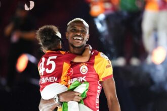 Two Galatasaray players celebrate, hugging on the field with smiles and a flag/cloth in hand.