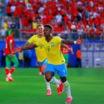 Brazilian soccer player in yellow jersey 7 celebrates with arms outstretched on a green pitch as teammates run by crowd in red in the stands behind him  trybalnews