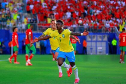 Brazilian soccer player in yellow jersey (#7) celebrates with arms outstretched on a green pitch as teammates run by, crowd in red in the stands behind him.