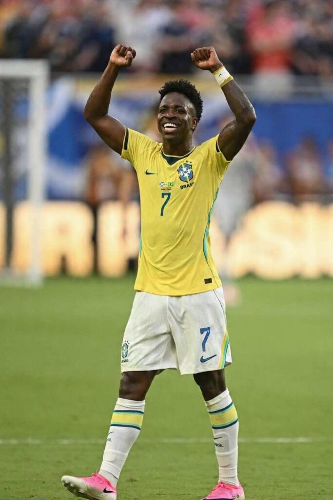 Brazil forward Vinicius Junior wearing yellow 7 jersey with green numbers and white shorts celebrates with both fists raised in the air He has a wide smile short curly hair and wears pink Nike boots with white socks with yellow green stripes Blurred stadium crowd and part of goalpost visible in background during FIFA World Cup 2026 Group C match between Scotland and Brazil at Miami Stadium  trybalnews