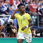 Brazil forward Vinicius Junior wearing yellow 7 jersey with green numbers and white shorts points to his temple with right hand in celebration gesture He has sweat on his face and looks toward the crowd with mouth open Blurred Scotland fans in blue and white plus photographers visible in background during FIFA World Cup 2026 Group C match between Scotland and Brazil at Miami Stadium Miami Gardens Florida on June 24 2026  trybalnews