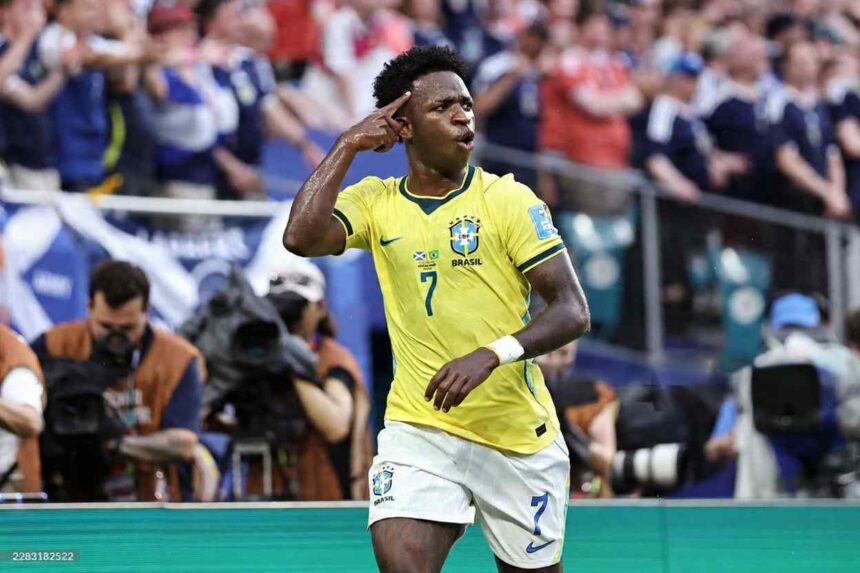 Brazil forward Vinicius Junior wearing yellow 7 jersey with green numbers and white shorts points to his temple with right hand in celebration gesture He has sweat on his face and looks toward the crowd with mouth open Blurred Scotland fans in blue and white plus photographers visible in background during FIFA World Cup 2026 Group C match between Scotland and Brazil at Miami Stadium Miami Gardens Florida on June 24 2026  trybalnews