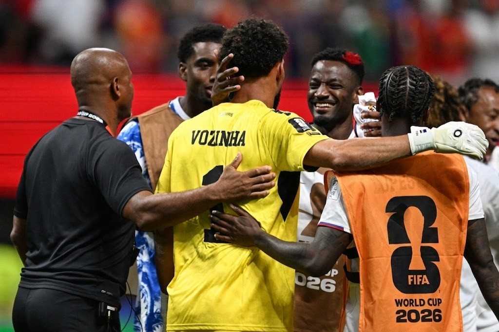 Vozinha Trybal News Soccer players celebrate in a group hug on the field one in a yellow jersey with the name VOZINHA visible on the back and a player wearing an orange bib with the FIFA World Cup 2026 logo on it trybalnews