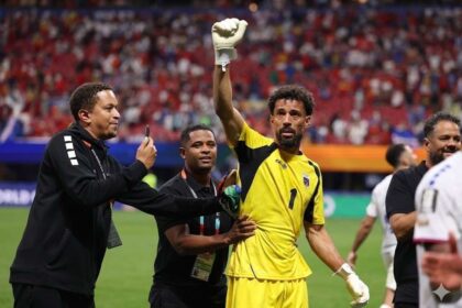 Football goalkeeper in a yellow jersey with number 1 (Vozinha) raises a gloved fist as teammates celebrate on the field.