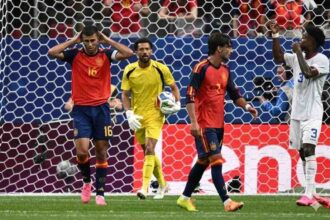 Soccer scene at the goalmouth: goalkeeper in yellow holding the ball, two players in red and blue nearby, and an opponent in white nearby, with the net in the background.