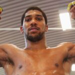 Male fighter with white hand wraps raises his clenched fists above his head in a gym sweat on his torso looking forward in a moment of victory  trybalnews