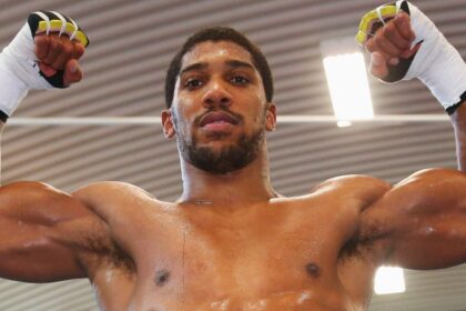 Male fighter with white hand wraps raises his clenched fists above his head in a gym, sweat on his torso, looking forward in a moment of victory.