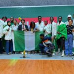 Group of athletes in white jackets posing with the Nigerian flag and a trophy on a gym floor  trybalnews