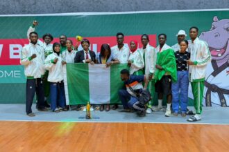 Group of athletes in white jackets posing with the Nigerian flag and a trophy on a gym floor.