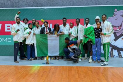 Group of athletes in white jackets posing with the Nigerian flag and a trophy on a gym floor.
