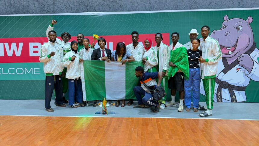 Group of athletes in white jackets posing with the Nigerian flag and a trophy on a gym floor  trybalnews