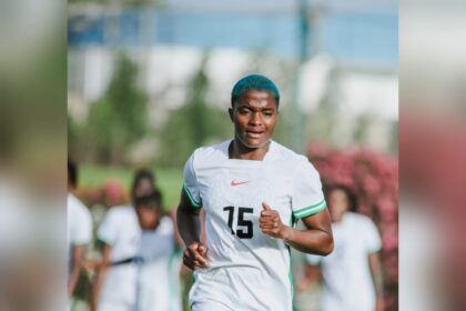 Soccer player with blue hair jogging on a field in a white kit with green trim, number 15 visible on jersey.