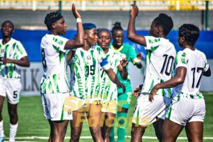 Group of female soccer players in white and green uniforms celebrate with raised arms on a green field after a goal or win.