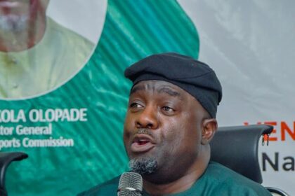 Man in green traditional attire speaks into a handheld microphone during a press conference, banners in the background.