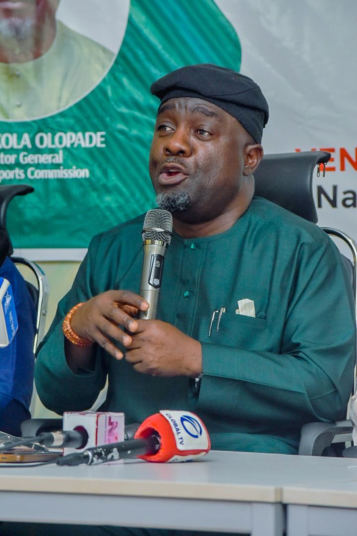 Man in green traditional attire speaks into a handheld microphone during a press conference banners in the background  trybalnews