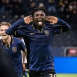 Soccer player celebrating a goal with hands cupping his ears and tongue out wearing a navy kit with a gold crest and number 36  trybalnews