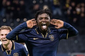 Soccer player celebrating a goal with hands cupping his ears and tongue out, wearing a navy kit with a gold crest and number 36.