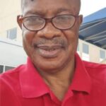 Portrait of an older man wearing glasses and a red polo shirt smiling slightly outdoors near a building  trybalnews