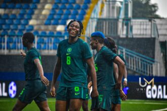 Soccer players in teal uniforms celebrate on a rainy field as stadium stands loom in the background.