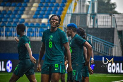 Soccer players in teal uniforms celebrate on a rainy field as stadium stands loom in the background.