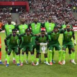 Nigeria national football team posing for a group photo on the pitch goalkeeper in orange on the left fans in stands behind them  trybalnews