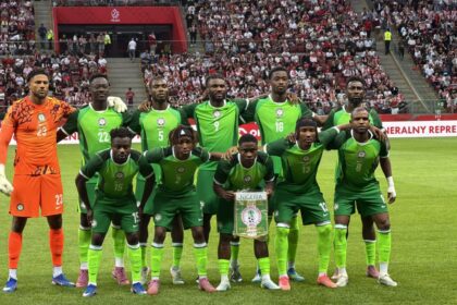 Nigeria national football team posing for a group photo on the pitch, goalkeeper in orange on the left, fans in stands behind them.