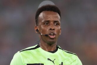 Soccer referee wearing a neon green kit with FIFA badge, headset mic, and black armbands, running on the field.