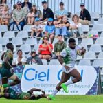 Rugby player in white and green runs with ball while a defender on the ground reaches to tackle crowd watches from stands behind a banner  trybalnews