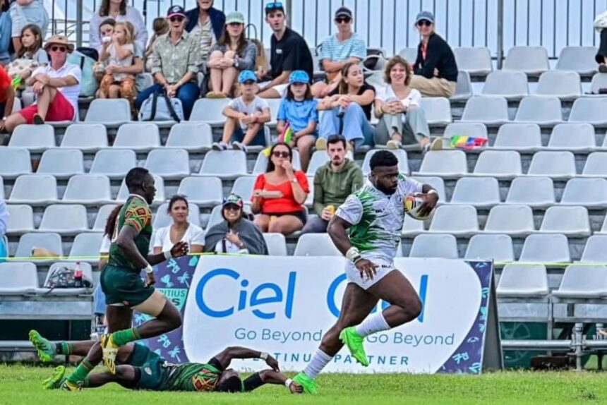 Rugby player in white and green runs with ball while a defender on the ground reaches to tackle crowd watches from stands behind a banner  trybalnews