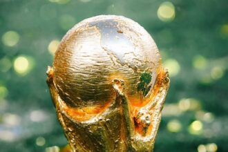 Golden World Cup trophy standing upright on green grass, close-up relief details visible on the sculpture.