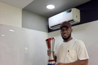 Man in a white t-shirt and maroon cap holds a tall silver trophy in a modern office setting.